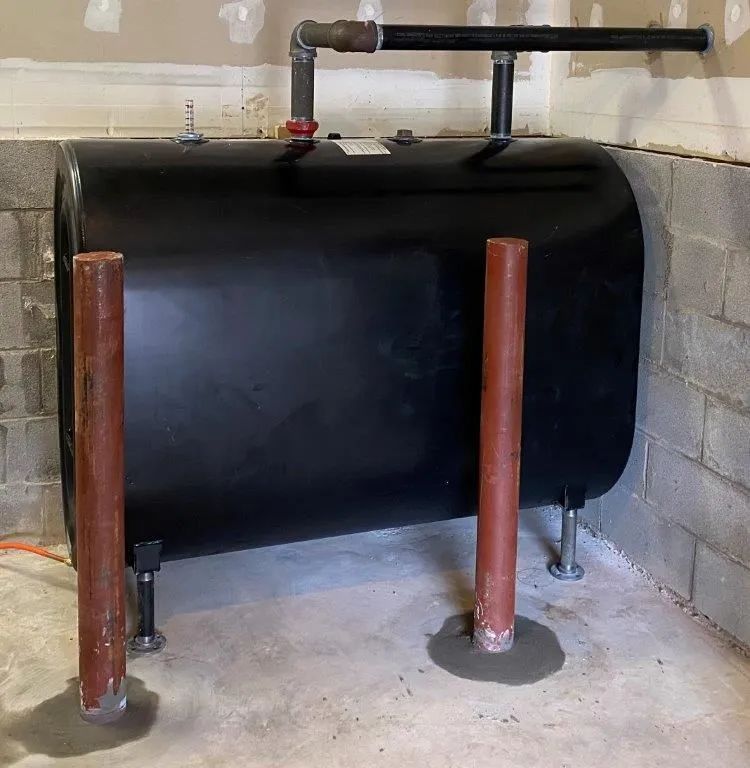 A black heating oil tank stands in a basement against concrete walls, protected by two red metal bollards.