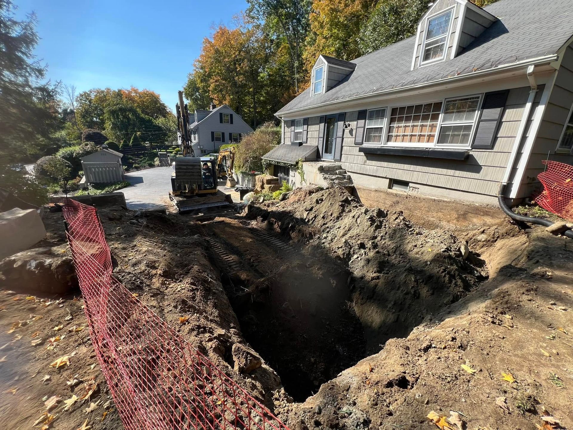 A construction excavator sits near a large open trench in front of a gray residential house on a sunny day.