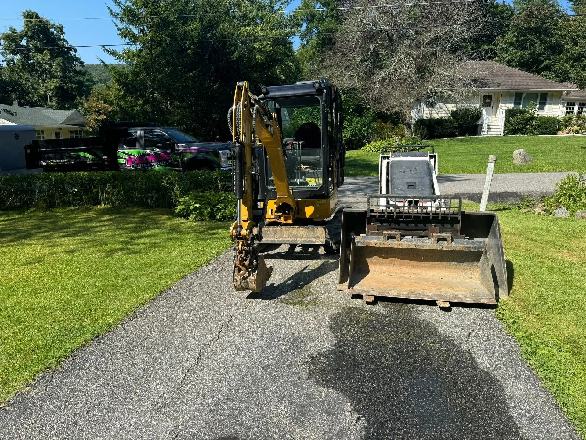 A yellow mini excavator and a skid steer bucket parked side-by-side on a gravel driveway in front of a house.