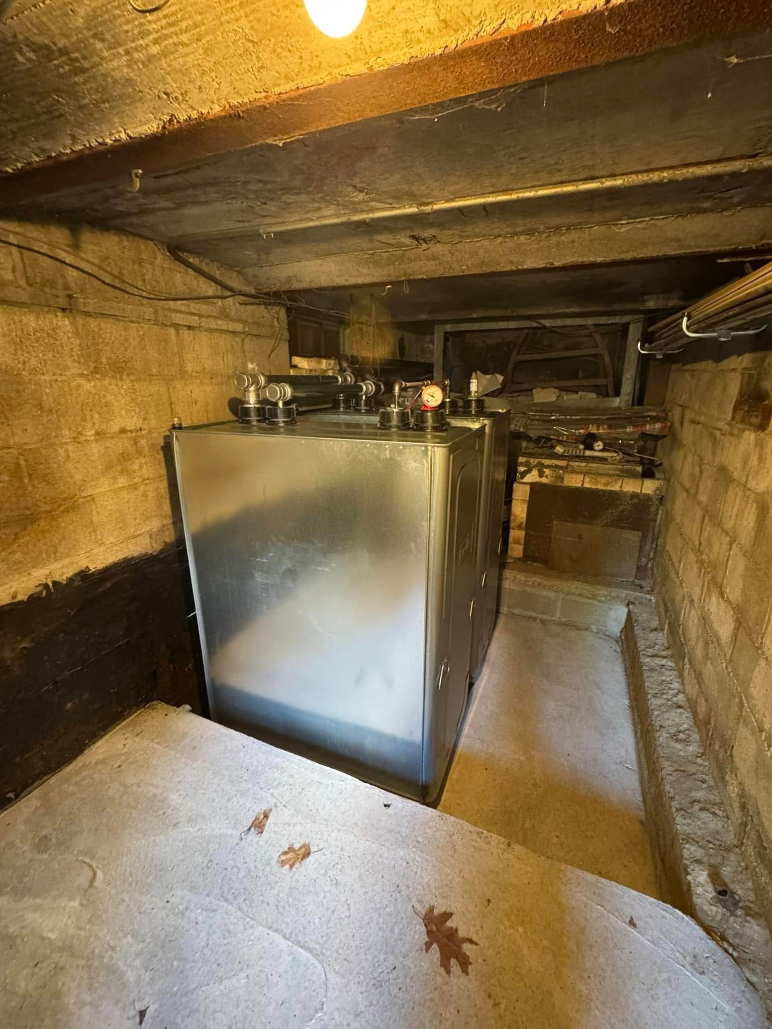 A large, rectangular silver metal storage tank positioned in a basement crawl space with stone walls and a concrete floor.