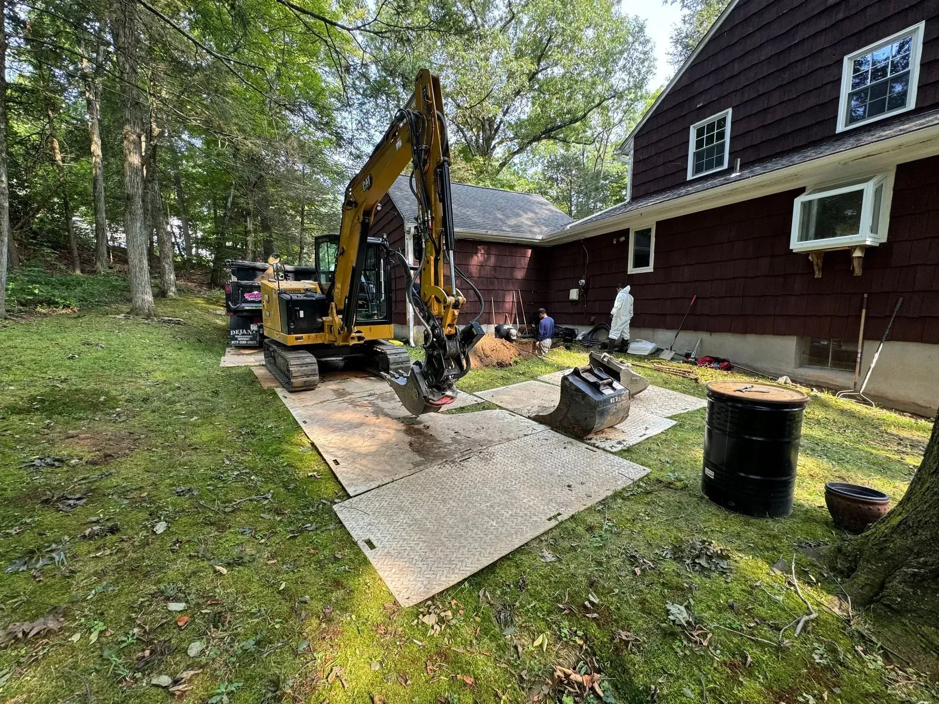 A yellow excavator operates next to a dark brown house, digging in the yard on temporary protective ground mats.