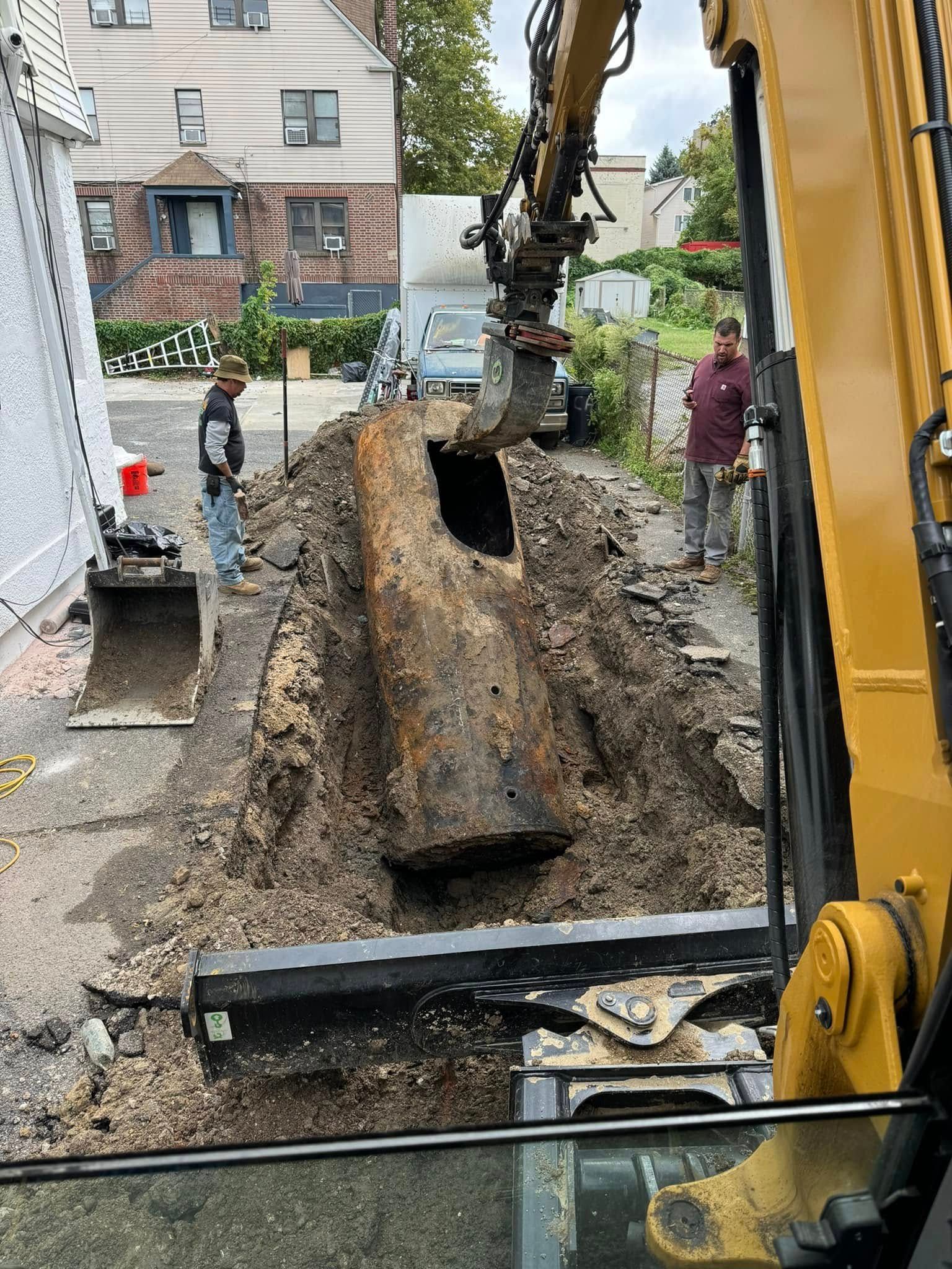 An excavator digs up an old, dark cylindrical tank buried in the ground while two people watch on a residential property.