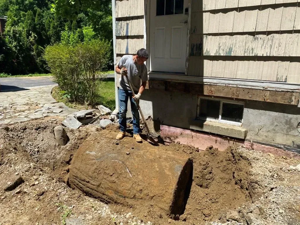 A person stands on a large, rusty, cylindrical underground storage tank partially unearthed next to a house foundation.