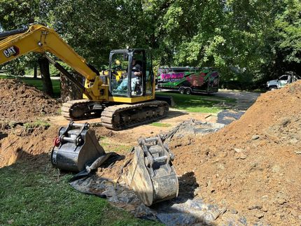 A yellow excavator bucket digging into wet soil near the foundation of a building.