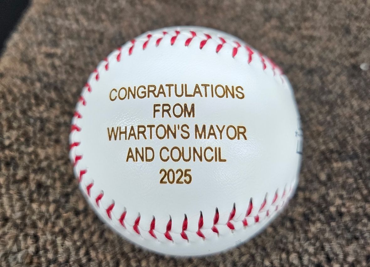 Baseball with gold text: CONGRATULATIONS FROM WHARTON'S MAYOR AND COUNCIL 2025 on a brown surface.