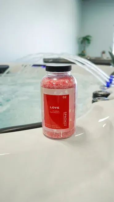Small red bottle with a black cap on a white surface in front of a blurred spa pool and plant