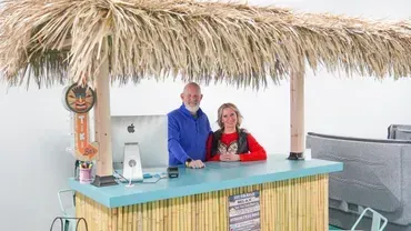 Two people stand behind a tiki-style bar under a thatched roof, smiling at the counter.