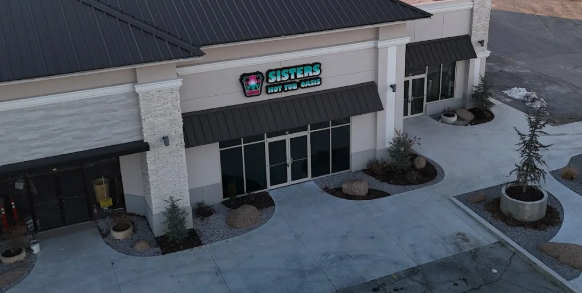 Front entrance of a building with glass doors, a lit sign, and landscaped planters by a curved walkway