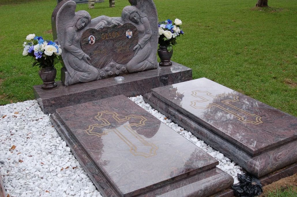 Grave site with a double headstone topped with angel statues; two rectangular flat markers. Green grass and white gravel surround.