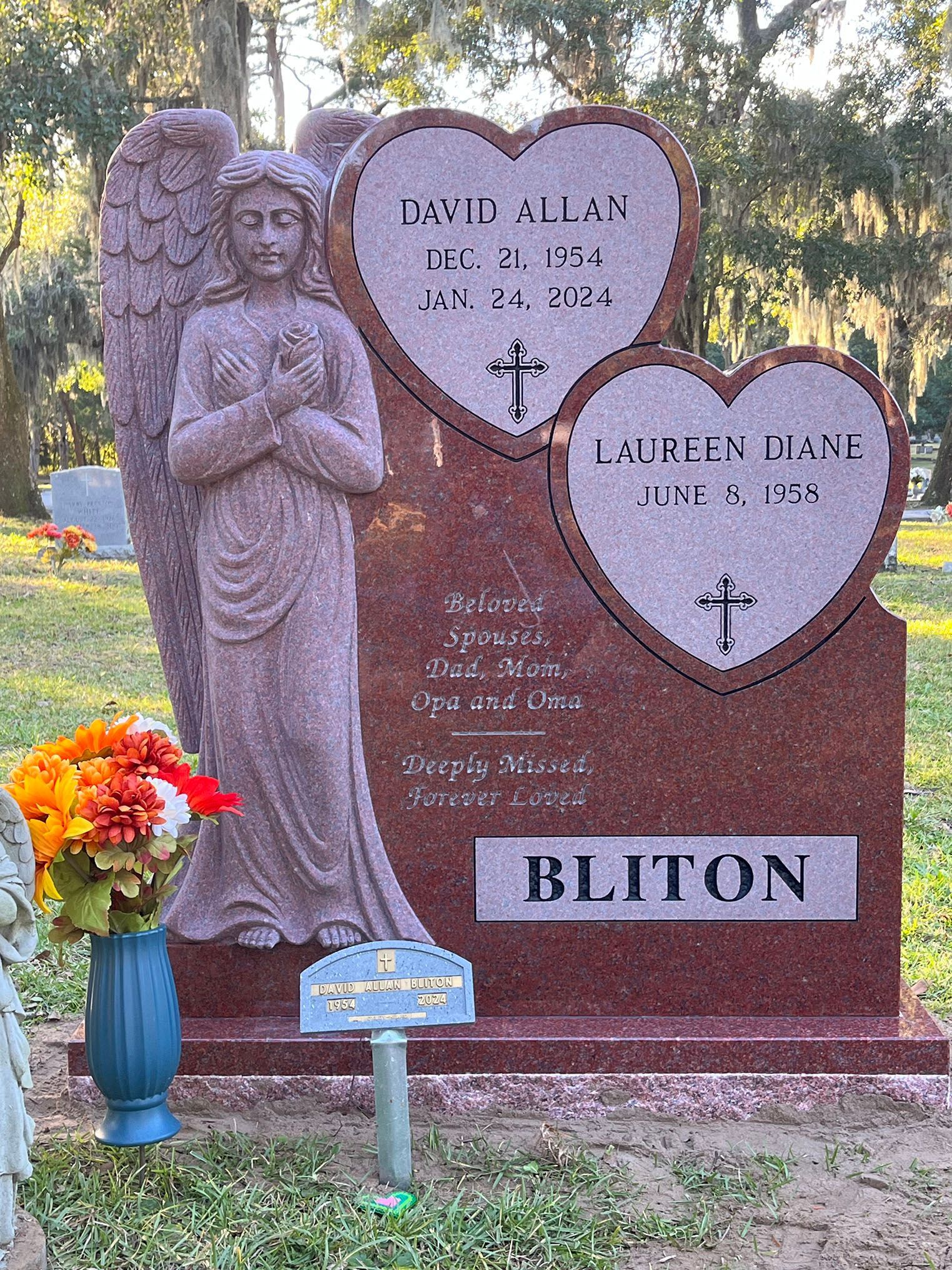 Red granite headstone with heart-shaped inscriptions, angel statue, and floral arrangement at a cemetery.