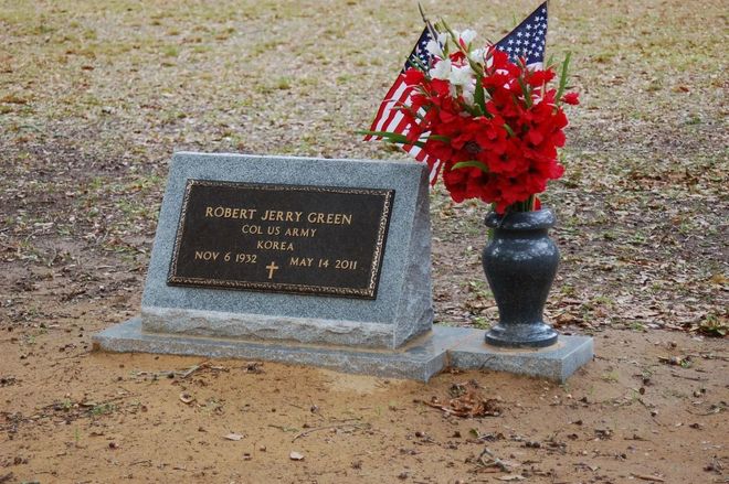 Grave marker with bronze plaque, flowers, and American flags in a cemetery.
