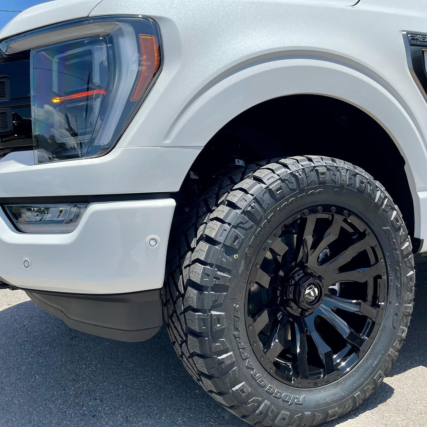 A white truck with black wheels and tires is parked on the side of the road.