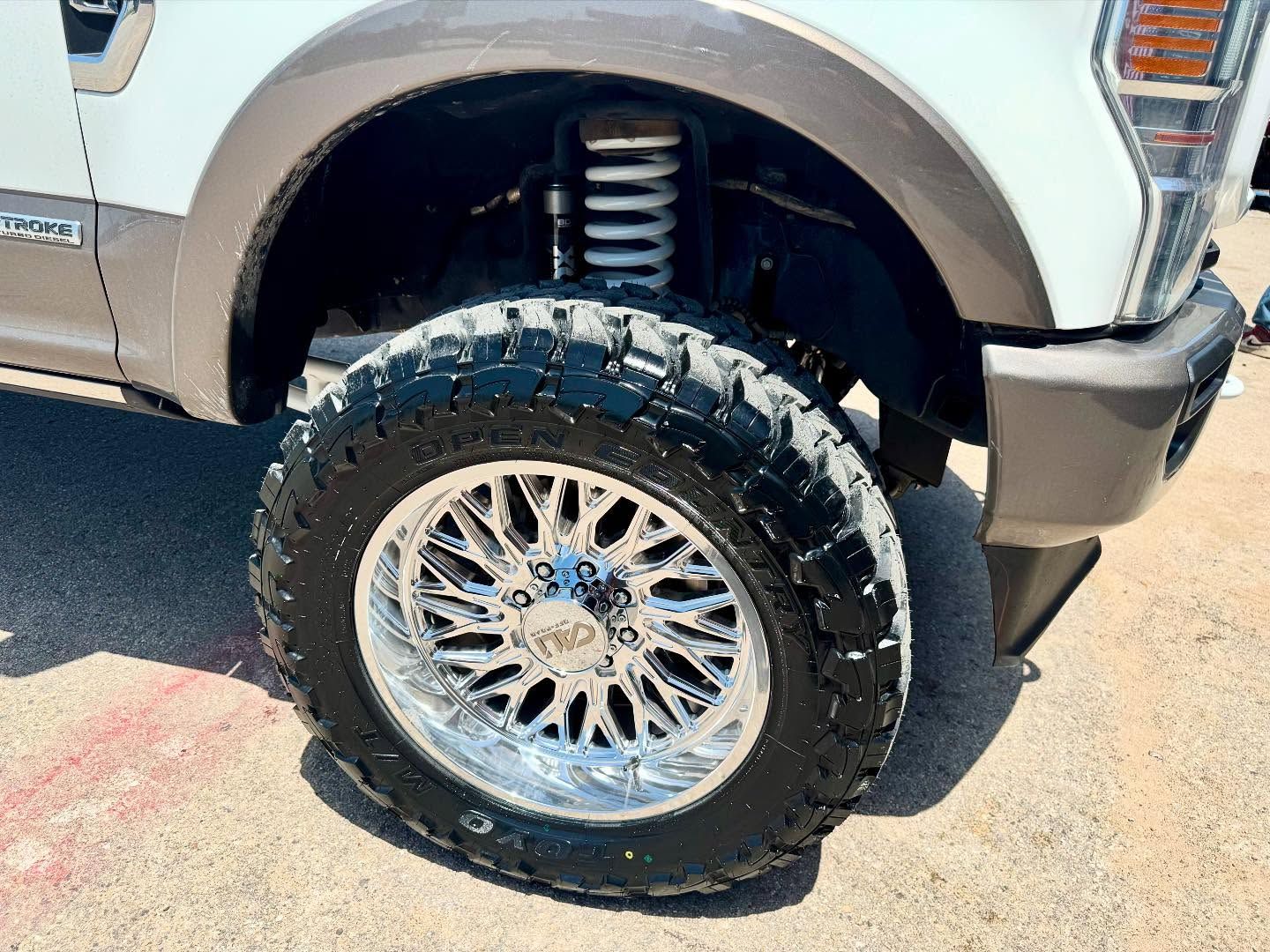 A close up of a white truck with a large tire on a dirt road.