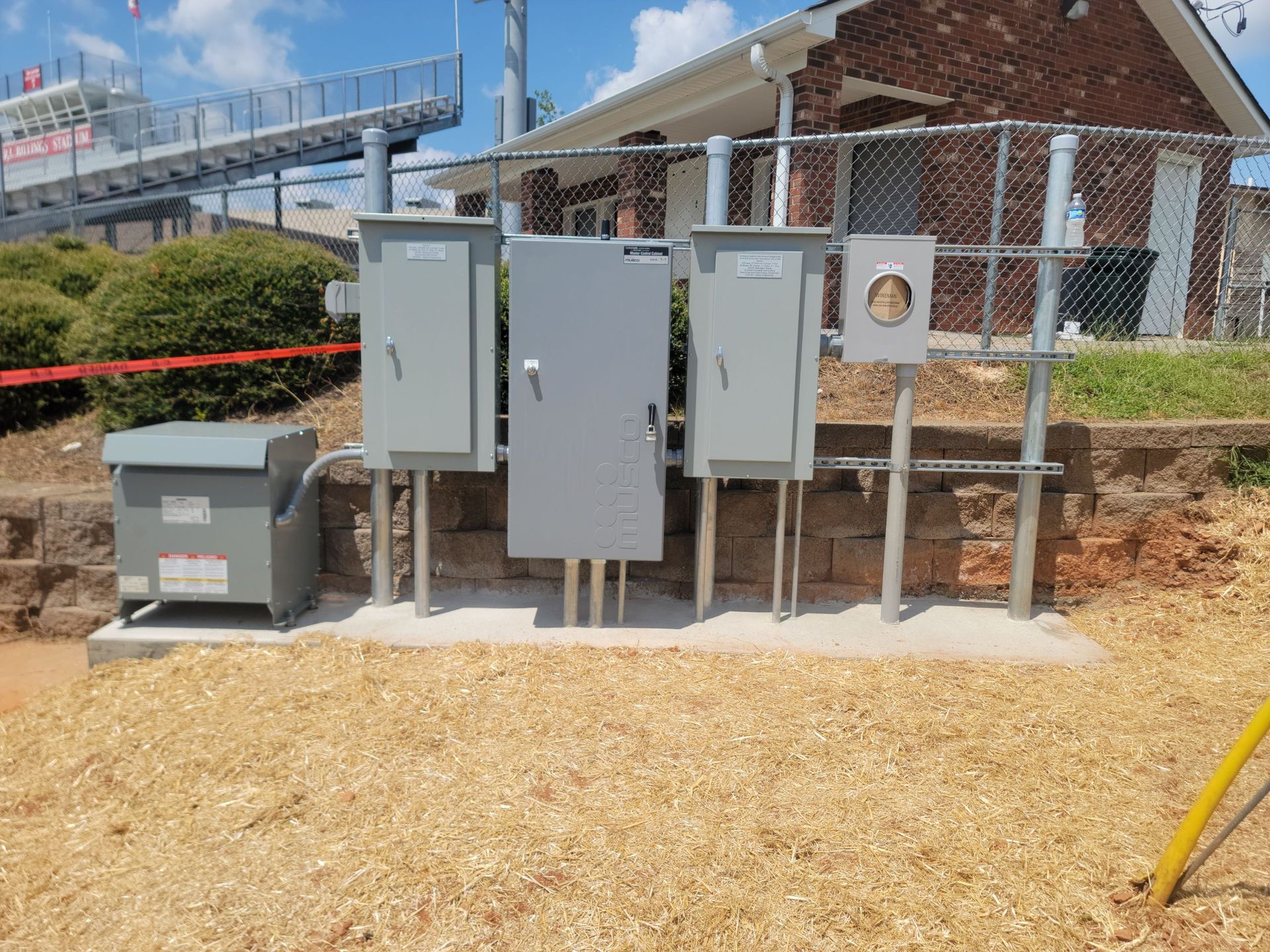 Gray electrical boxes and transformer on concrete base near a fence and building, on wood chips.