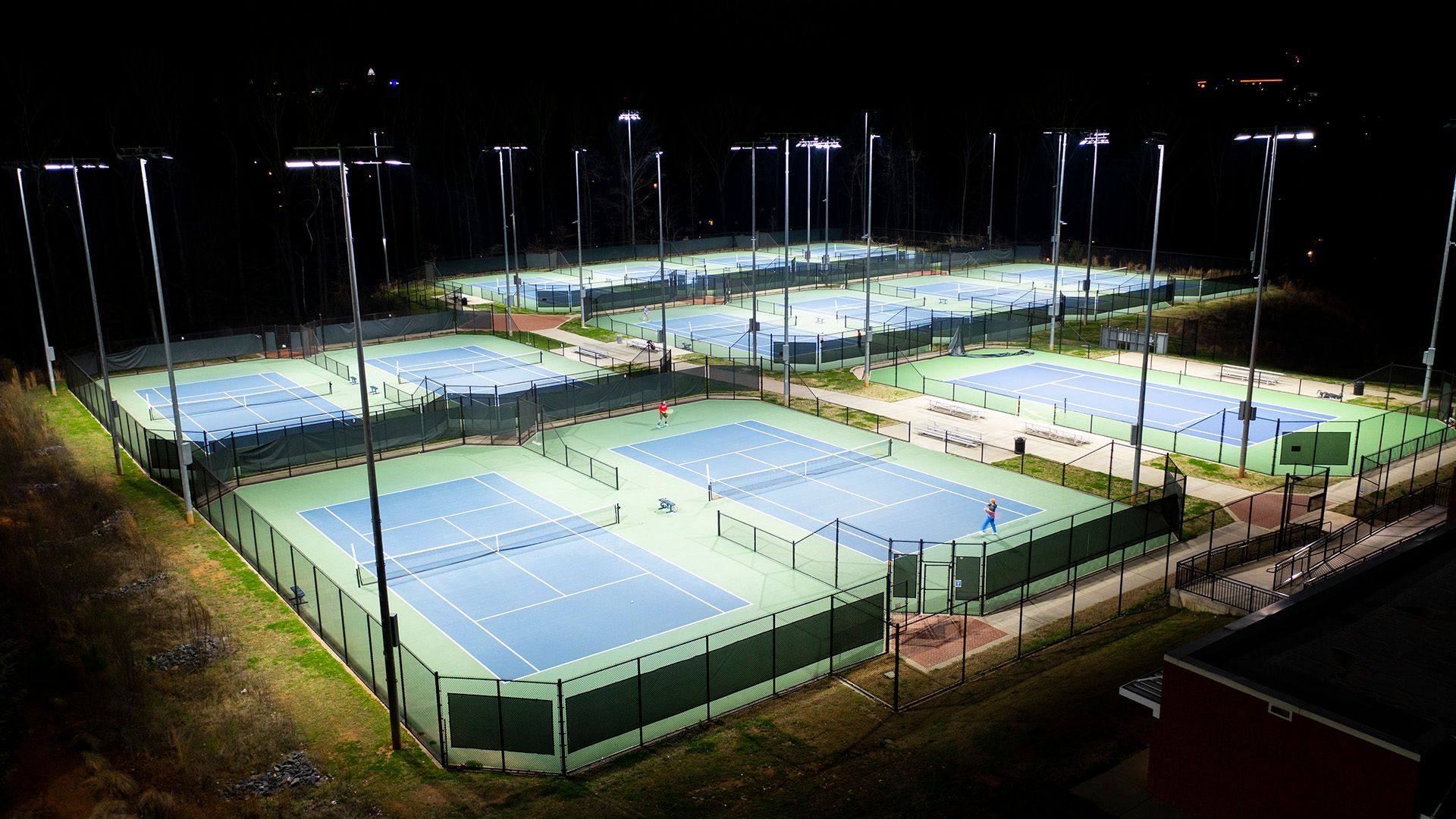 Lighted tennis courts at night. Several blue-green courts are surrounded by fences. Tall light poles illuminate the area.