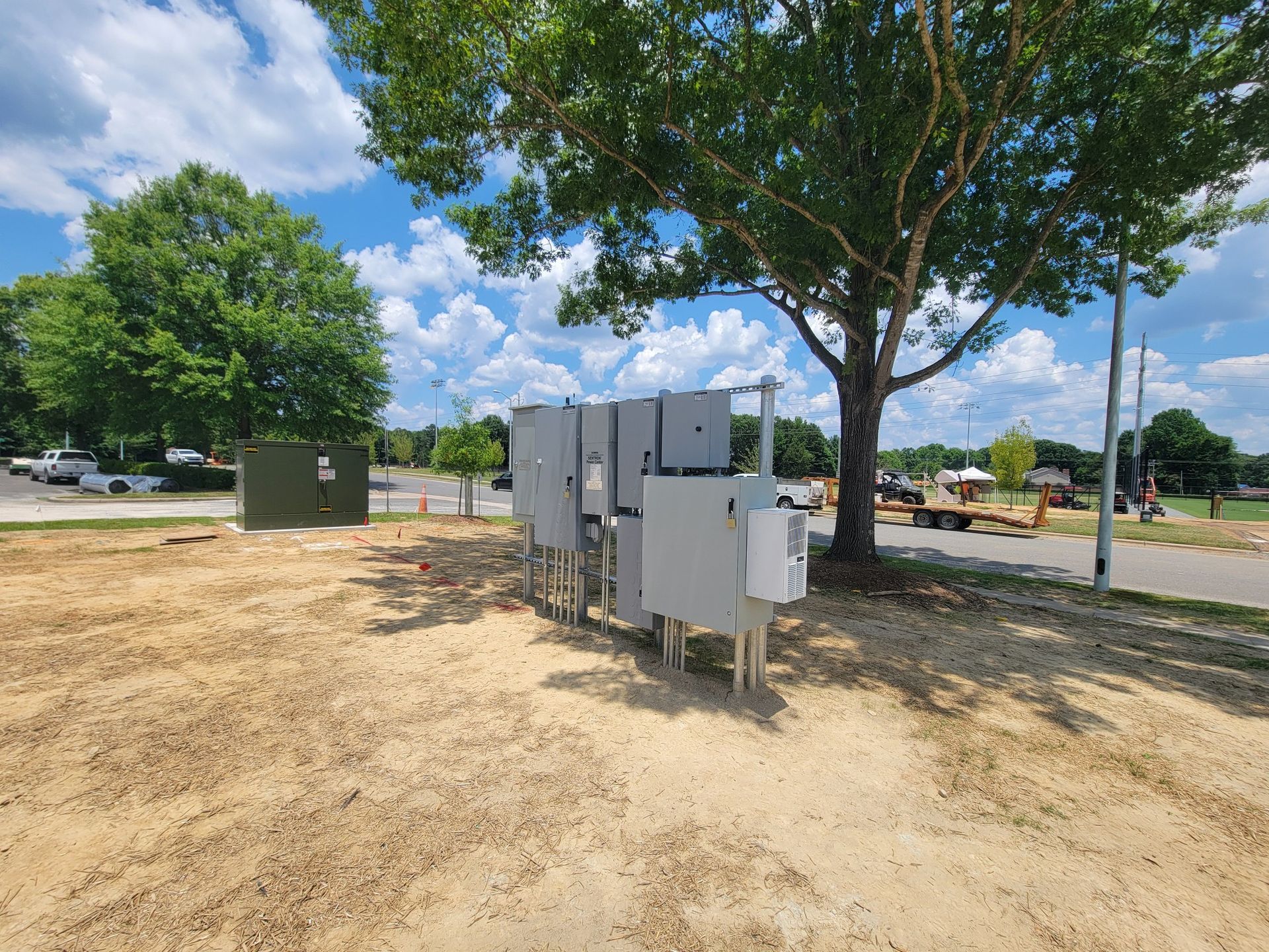 Electrical equipment boxes on a dirt patch under a tree, with a construction site in the background.