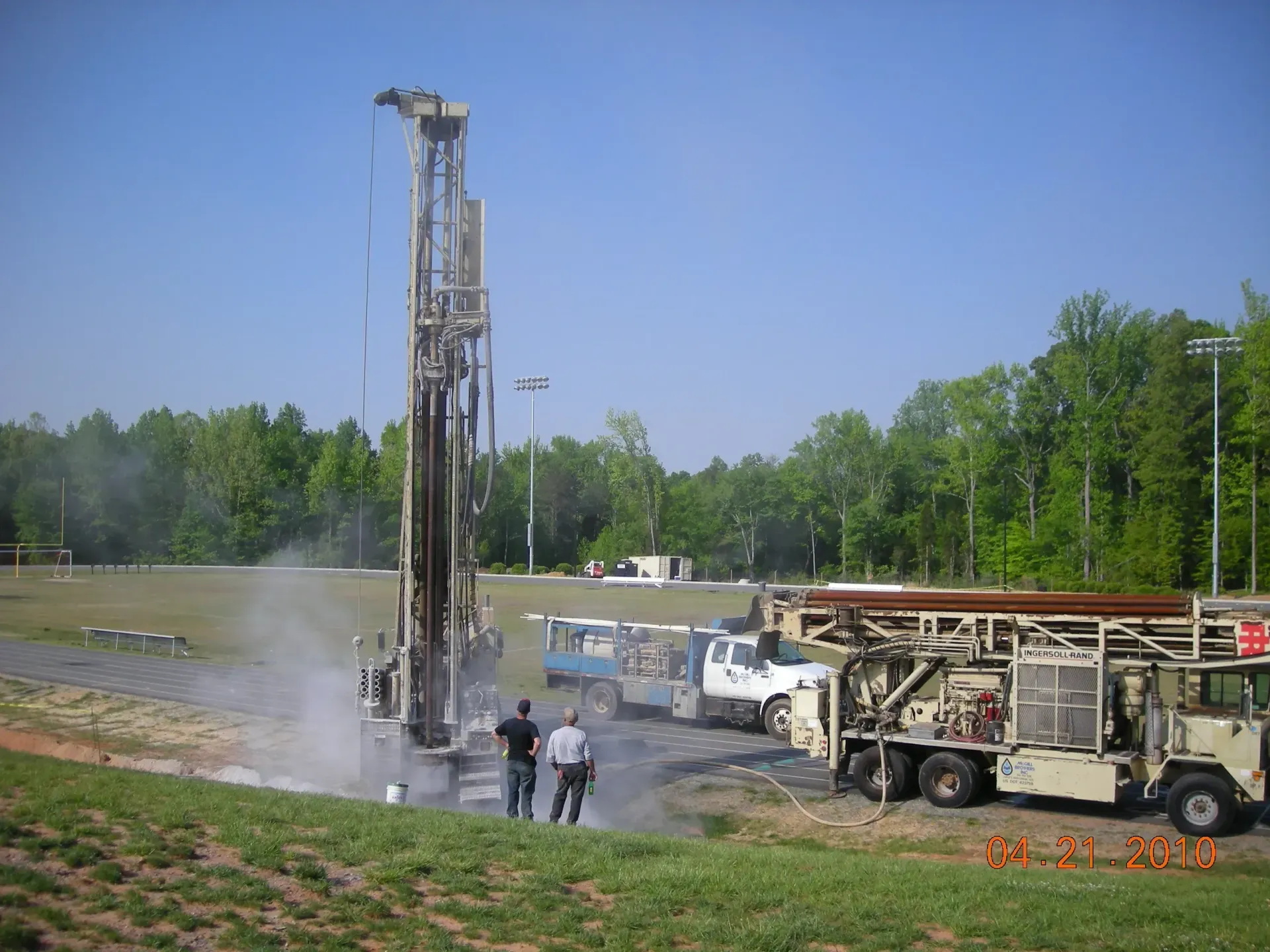 Drilling rig in a field, two people watching. Water spraying. Trucks and trees visible.