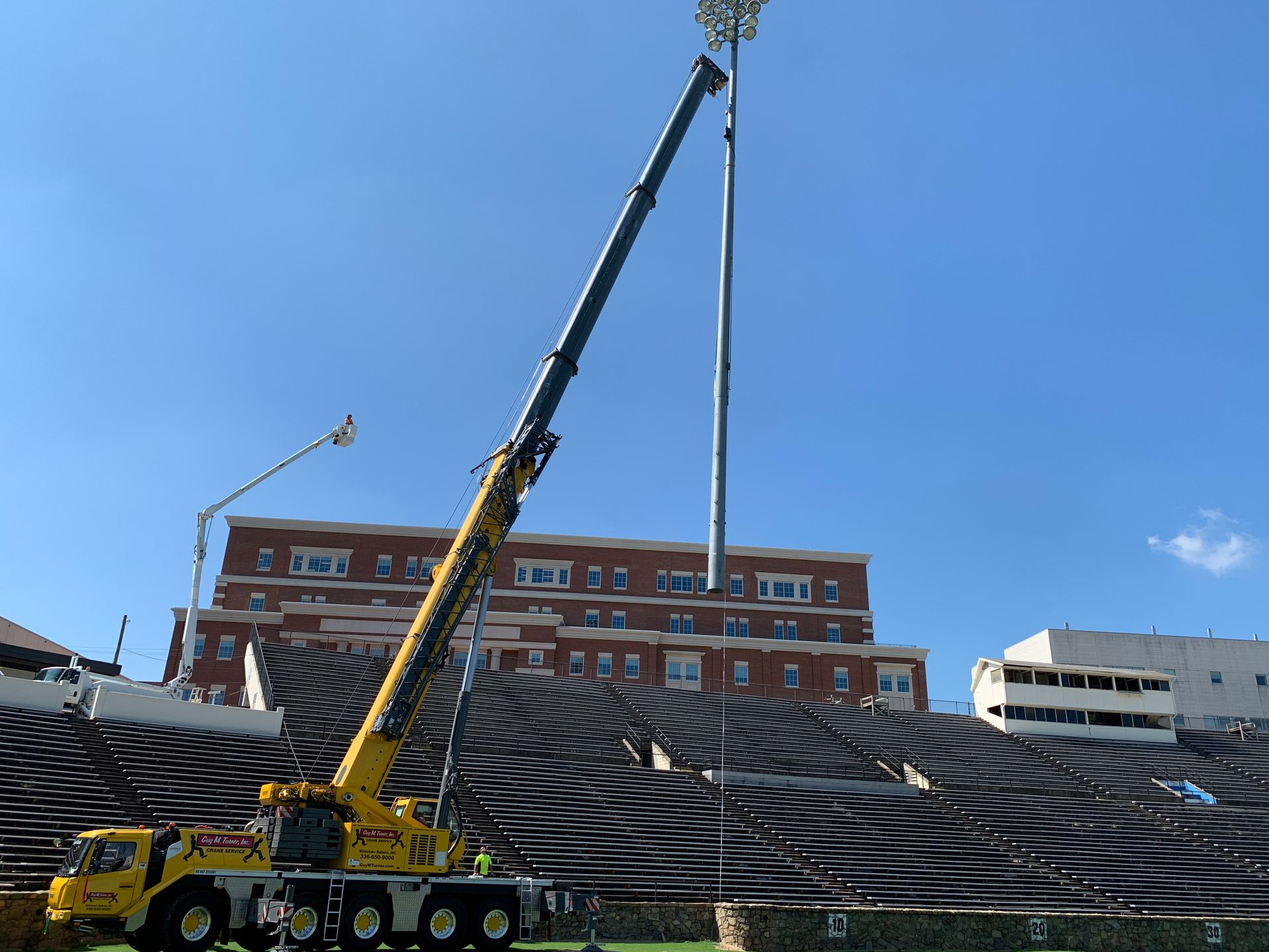 A large yellow crane lifting a light pole above a stadium, blue sky backdrop.