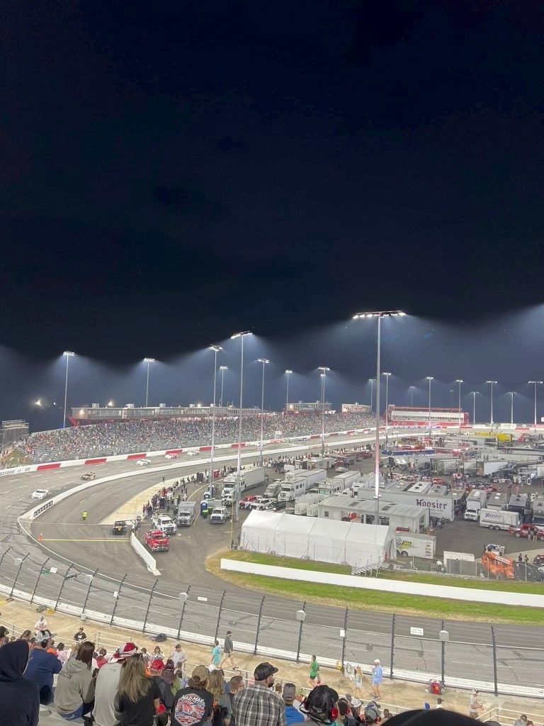 Night view of a racetrack with a packed grandstand. Race cars on track, with many trailers in the infield.