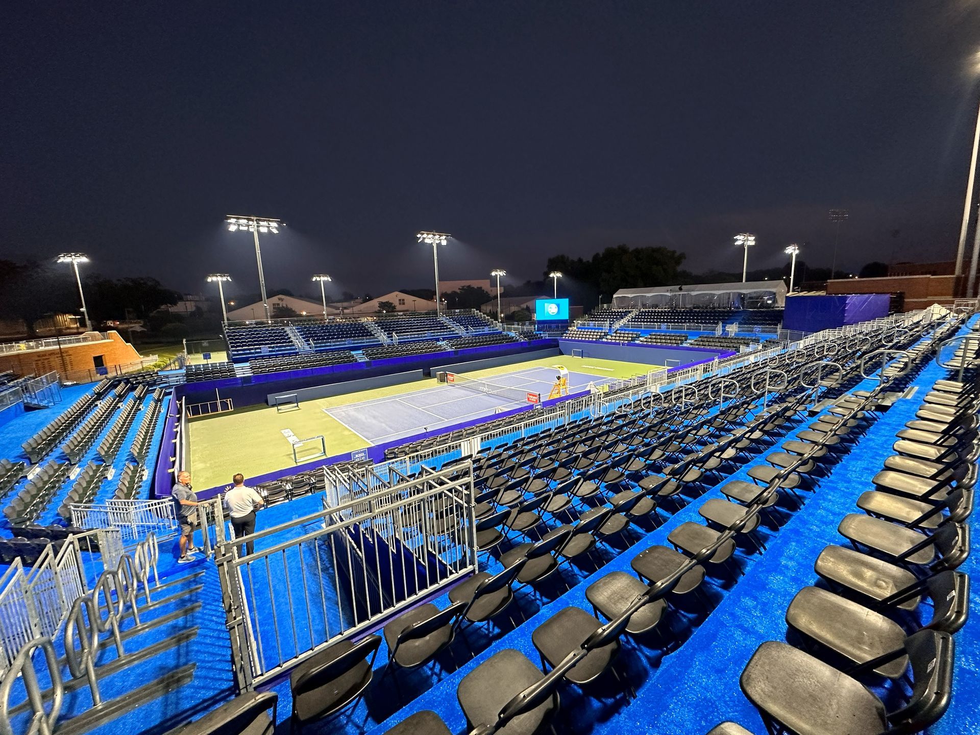 Tennis court at night with empty blue and black seating.