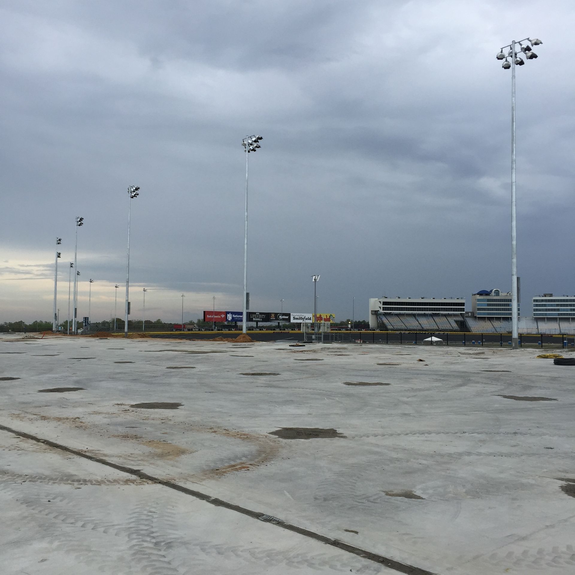Empty concrete lot with tall light poles under a cloudy sky. Buildings and freight trains in the background.
