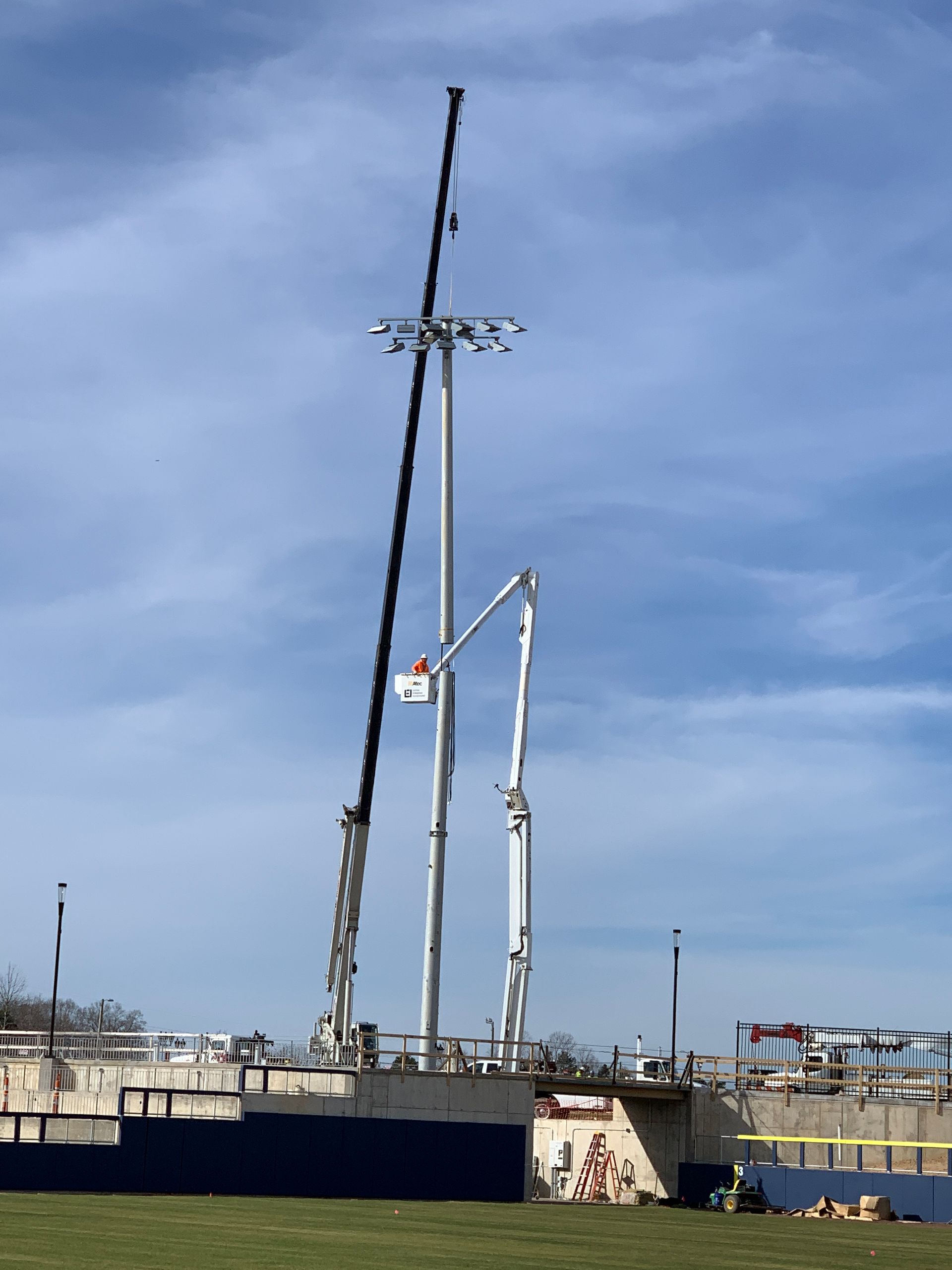 Construction of stadium lights; crane and lift platform are used. Blue sky, green grass, and stadium seating are also visible.