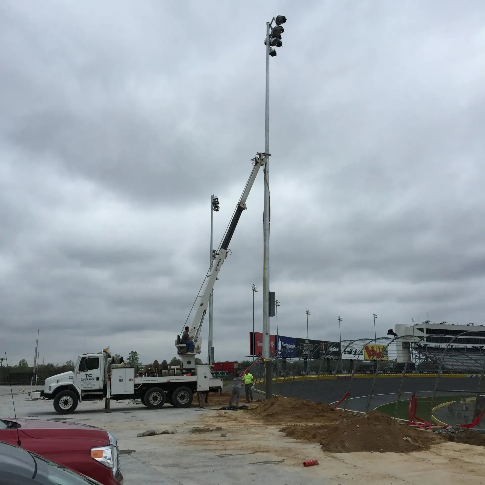 Truck with lift arm installing a tall light pole at a racetrack, under a cloudy sky.