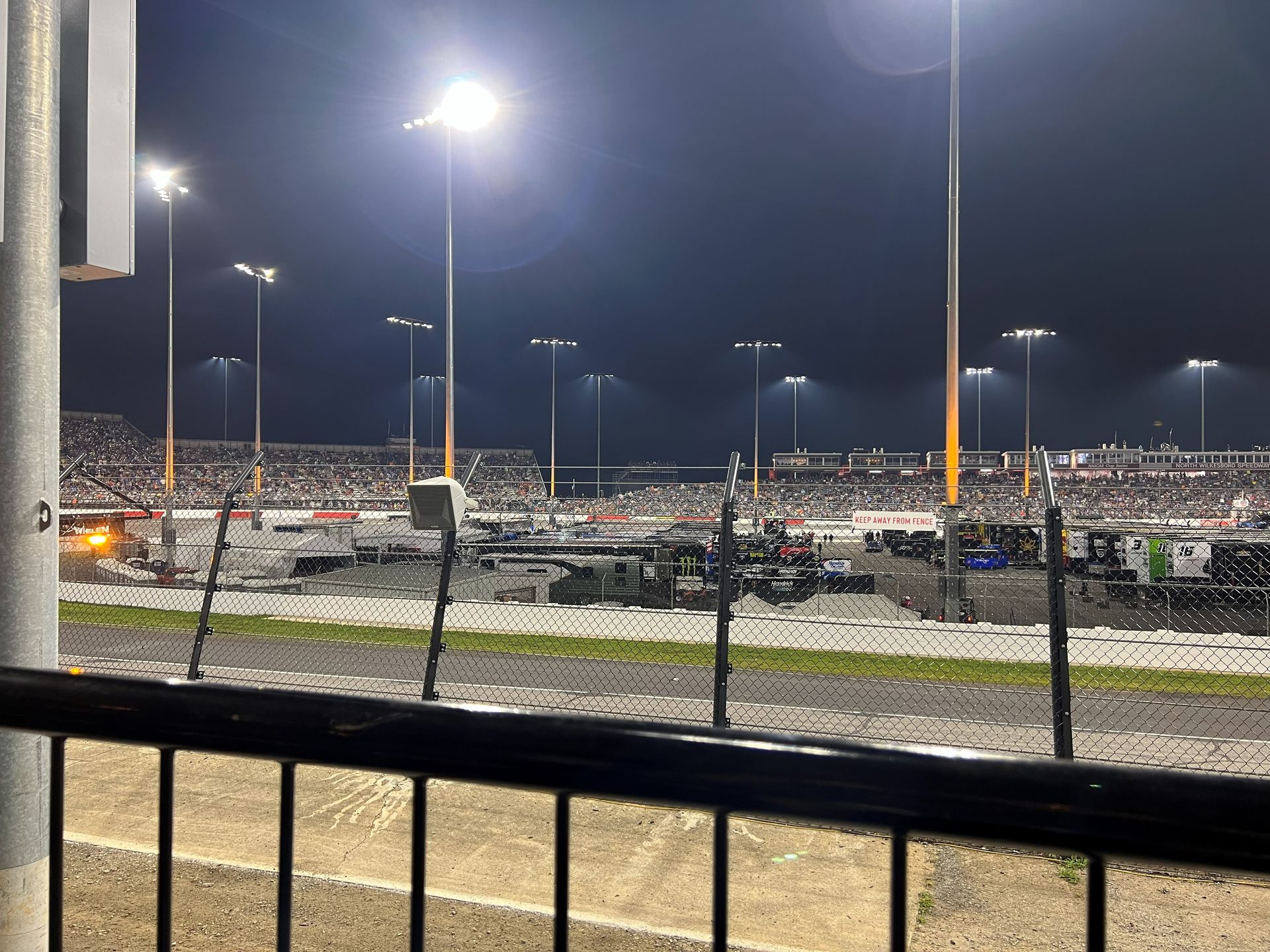 Nighttime race track scene with stadium lights illuminating a crowd, fence in foreground.
