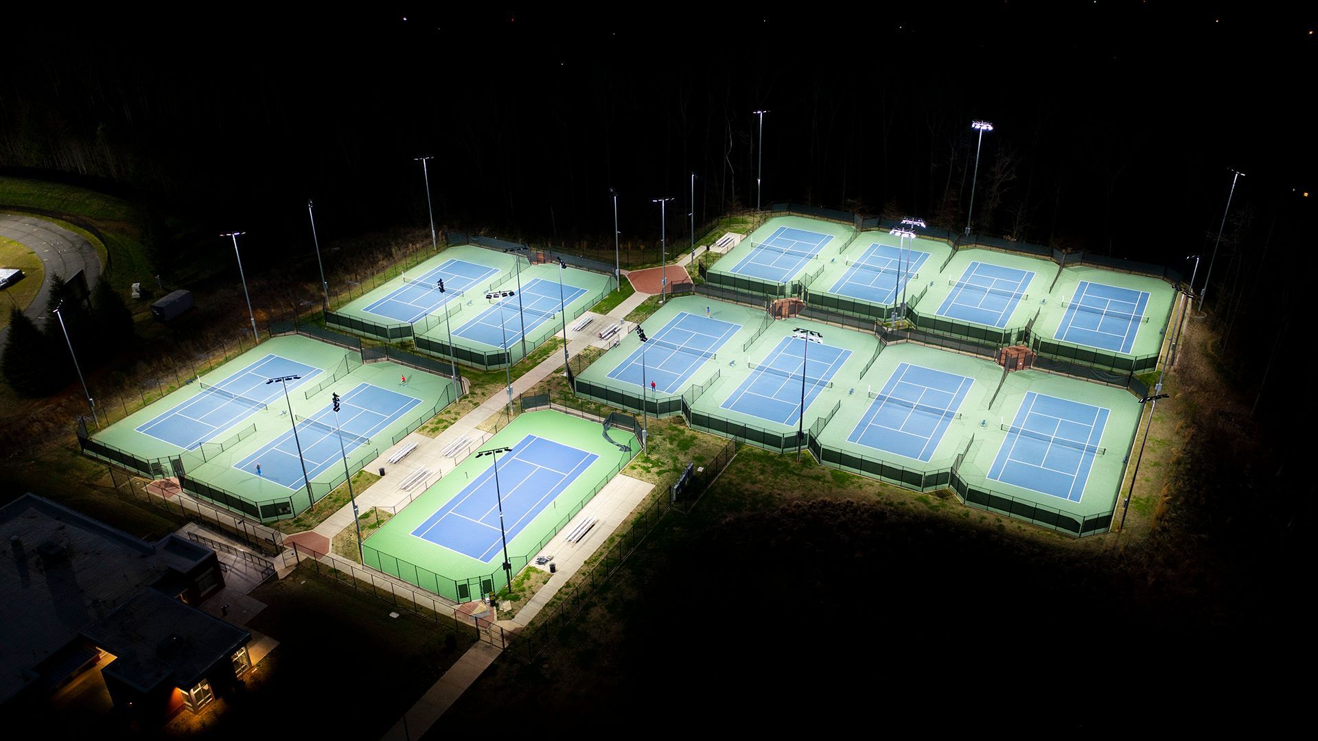 Lit pickleball courts at night. Blue courts with players and bright overhead lights.