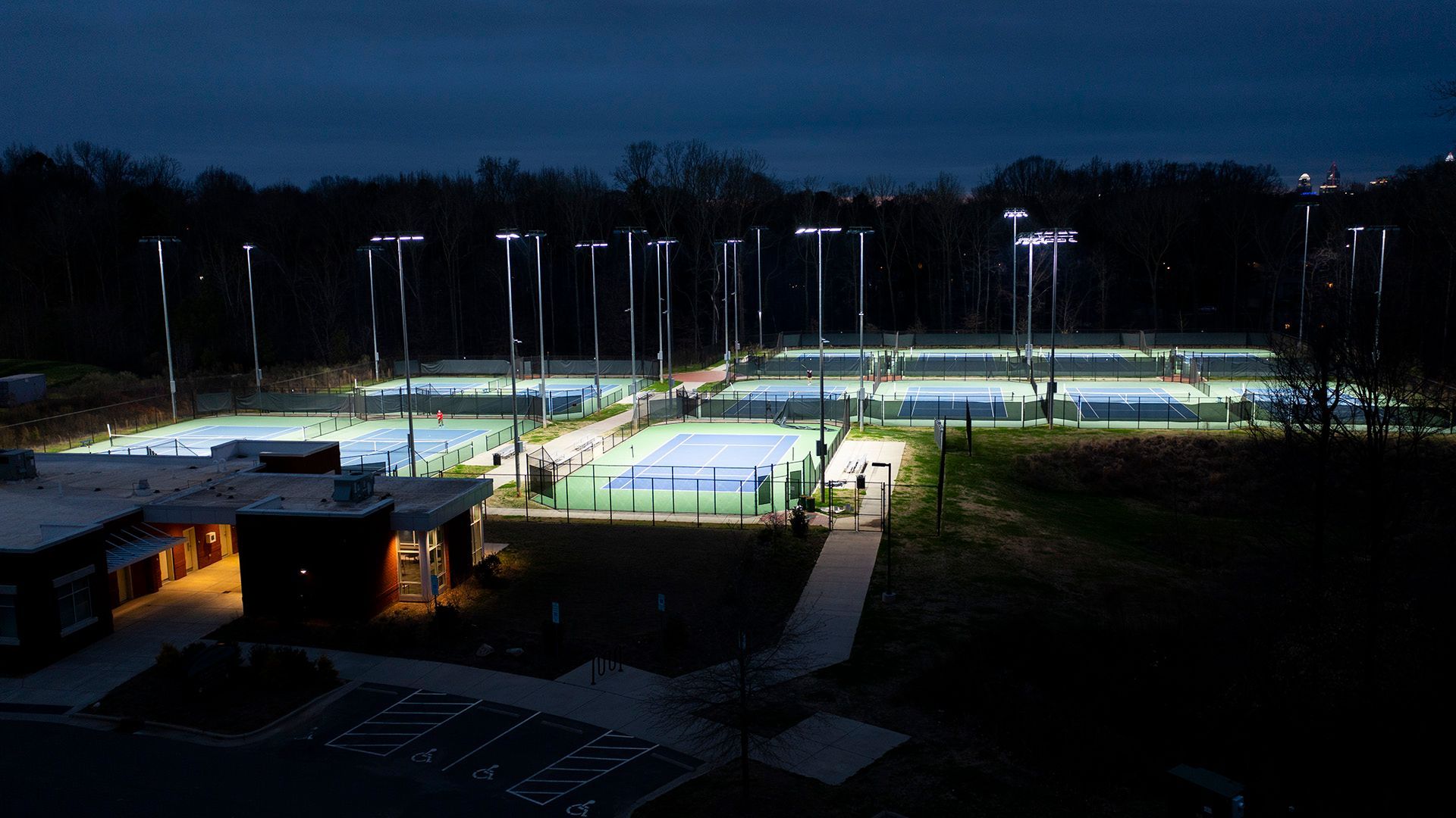 Lit tennis courts at night with a building in the foreground and trees in the background.
