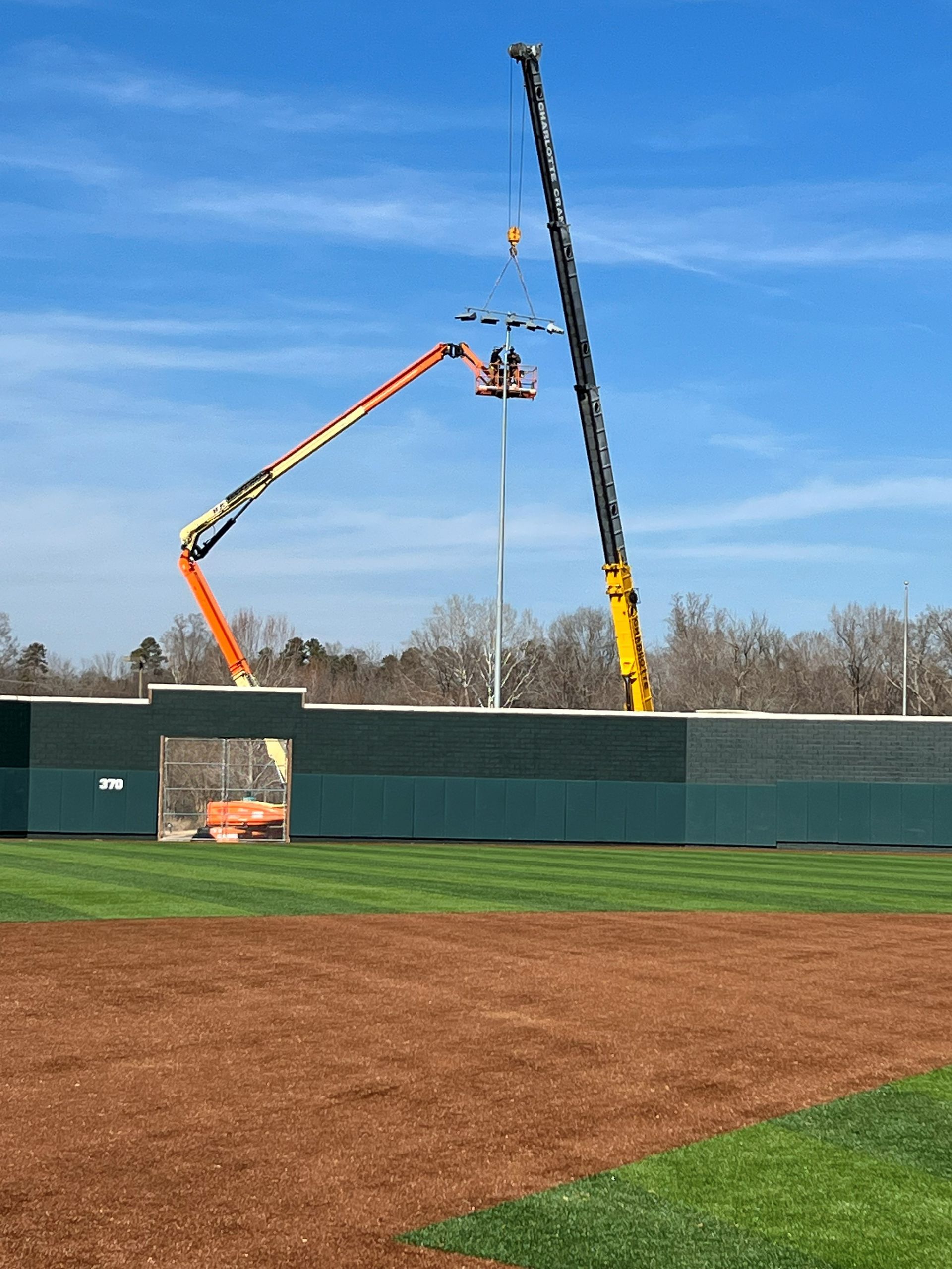 Two cranes working on a baseball field's light pole, against a blue sky. One is orange, the other is yellow.