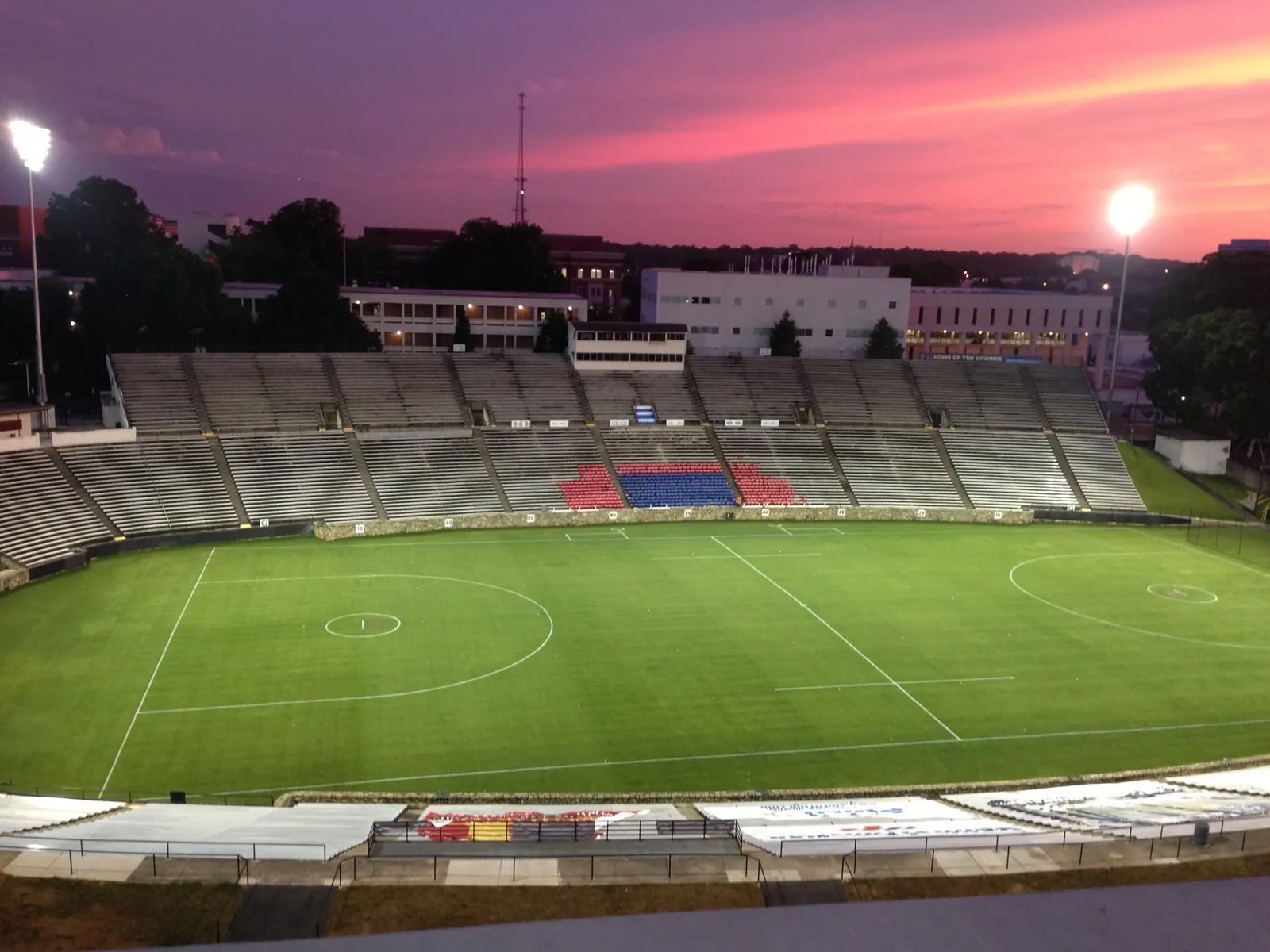 Stadium with green field, empty concrete seating, and a pink and purple sunset.