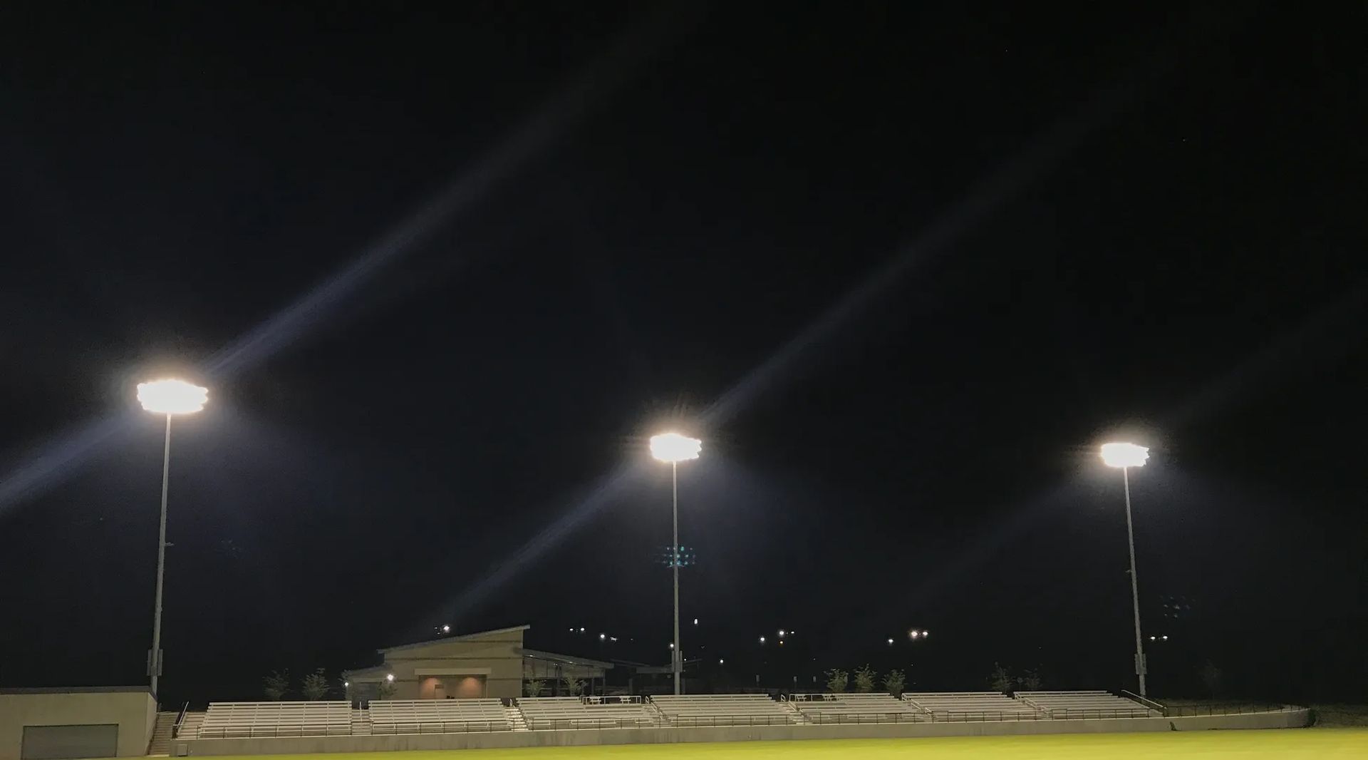 Nighttime view of a sports field with illuminated stadium lights and a distant building.