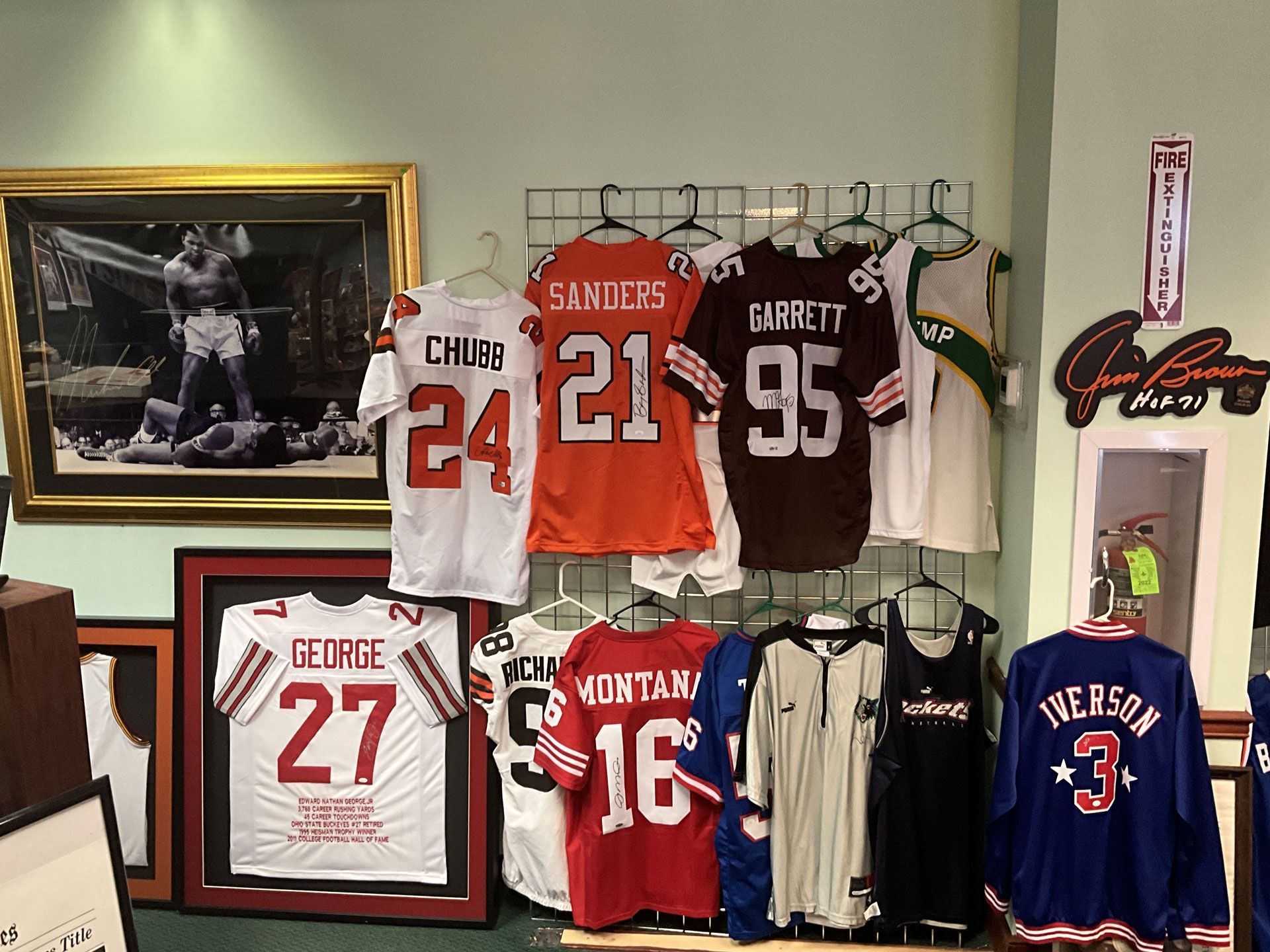 Sports jerseys hanging on a wall, framed photos, and a sign. Various colors and numbers visible.
