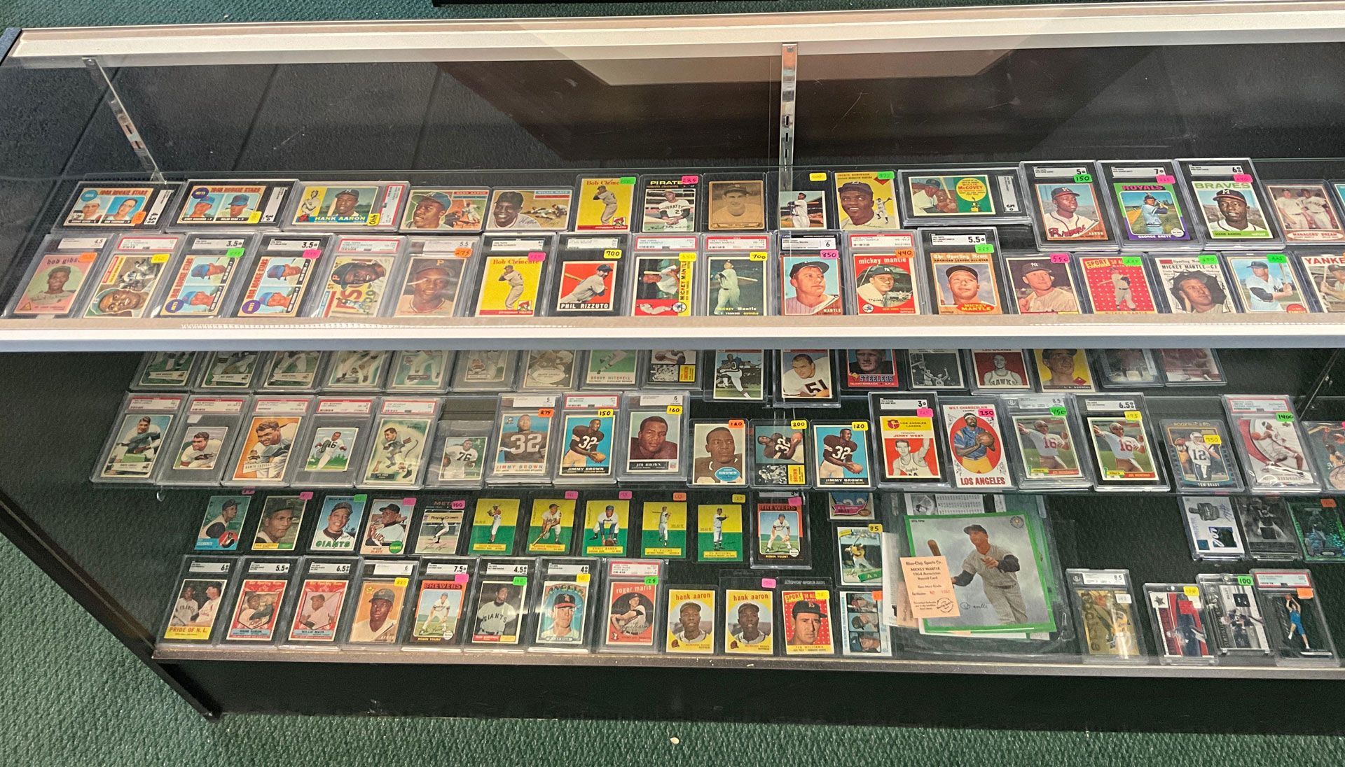Baseball cards displayed in a glass cabinet. Cards are in protective holders, various colors, store setting.