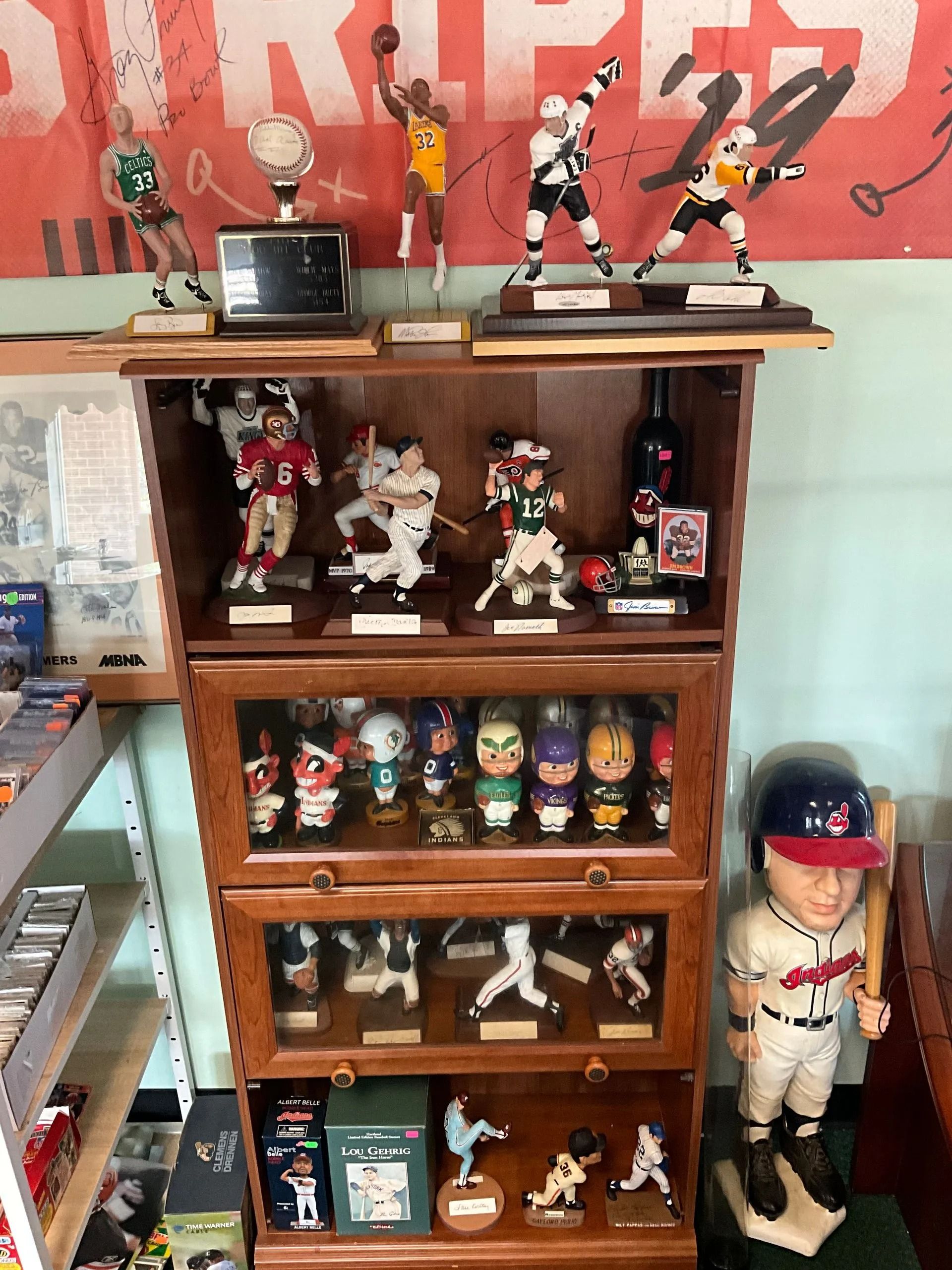 Wooden display case filled with sports figurines and bobbleheads. A large baseball player statue stands next to it.
