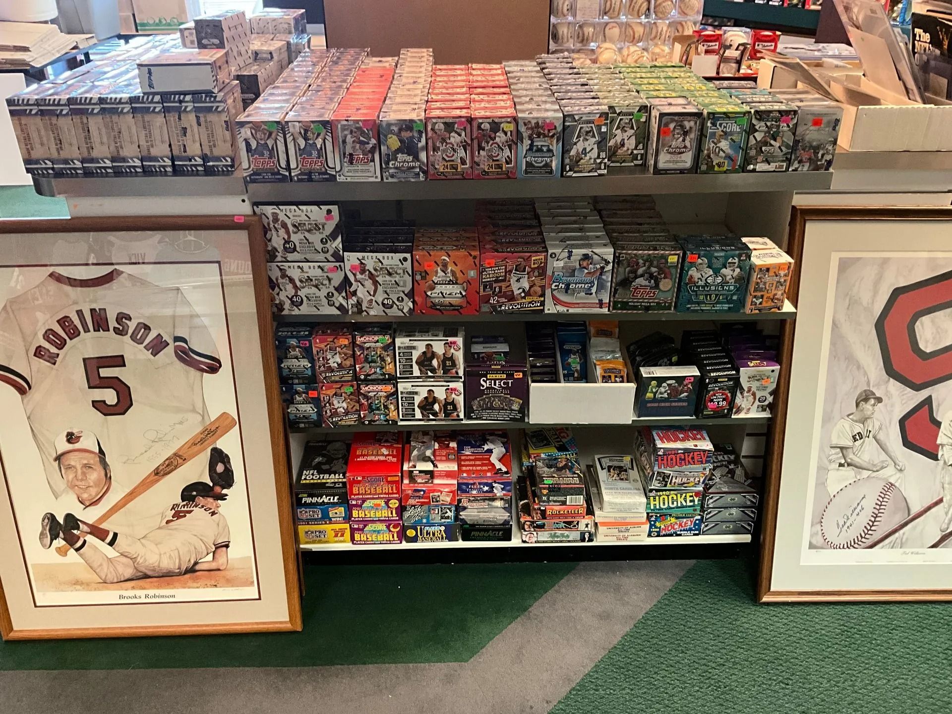 Shelves displaying trading card boxes, framed baseball jersey and poster, green carpet.