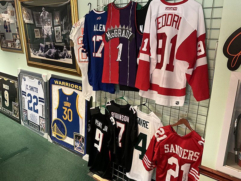Sports jerseys, including Raptors, Red Wings, and Sanders, hang on a wire rack. Framed memorabilia on the left.