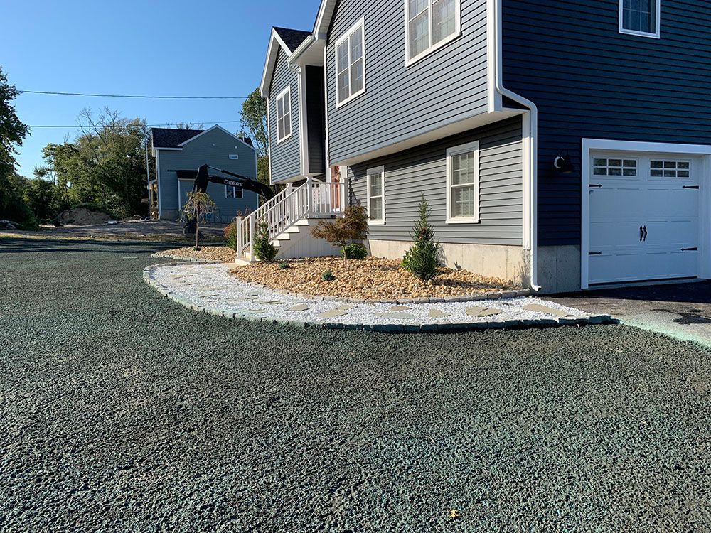 A landscape featuring gravel driveway, rock wall, and trees in front of a house.
