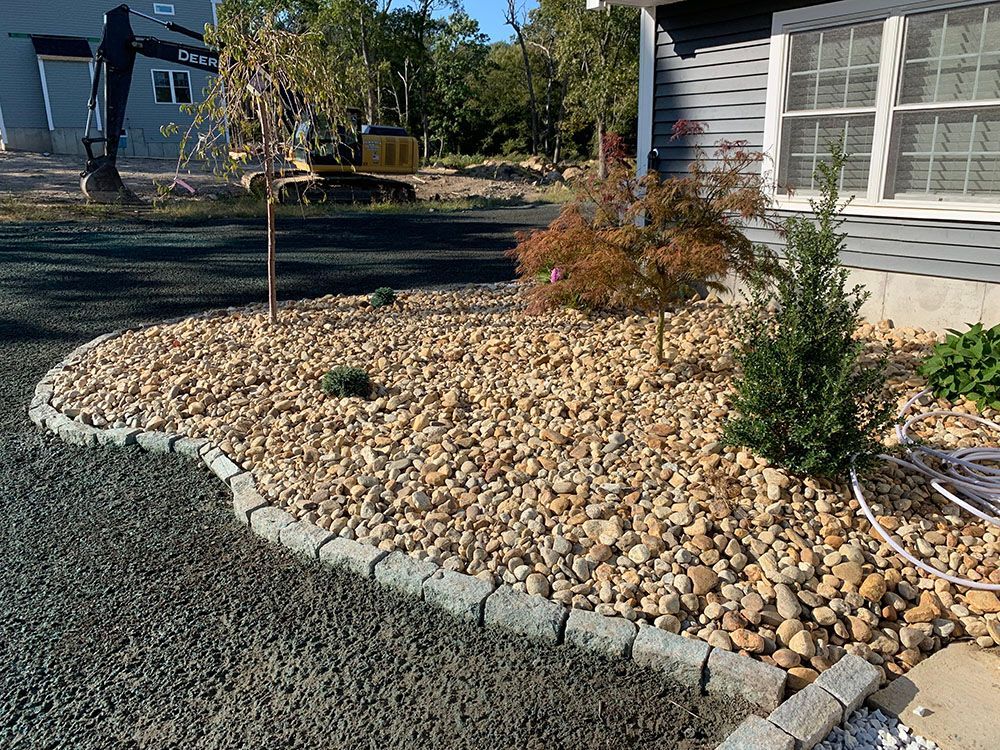 Gravel driveway with rock wall and tree in background.
