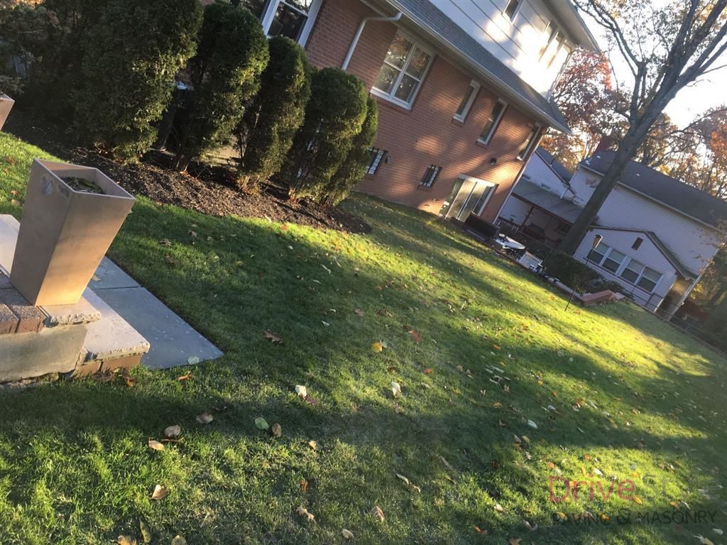 Tilted view of a grassy yard beside a brick house, with trees, shrubs, and a small concrete structure in the foreground