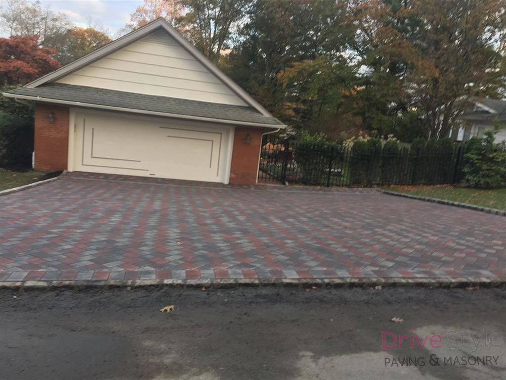 Brick-paved driveway in front of a beige garage, with trees and a fence in the background