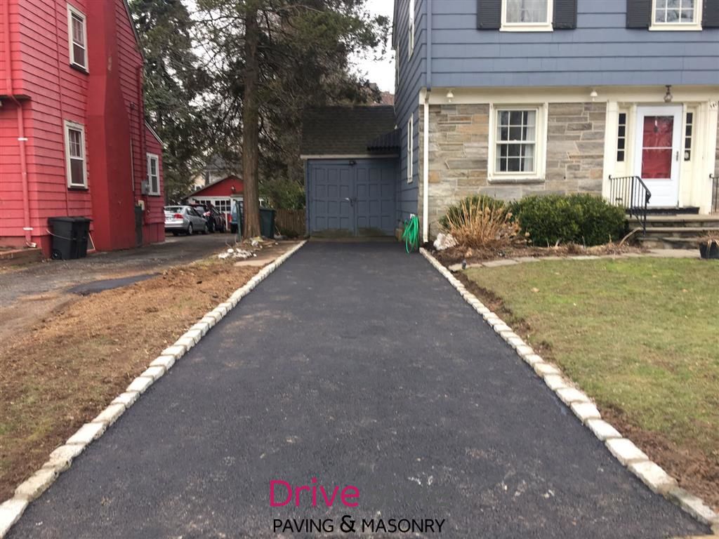 Freshly paved black asphalt driveway between red and gray houses, leading to a garage with a green bin.