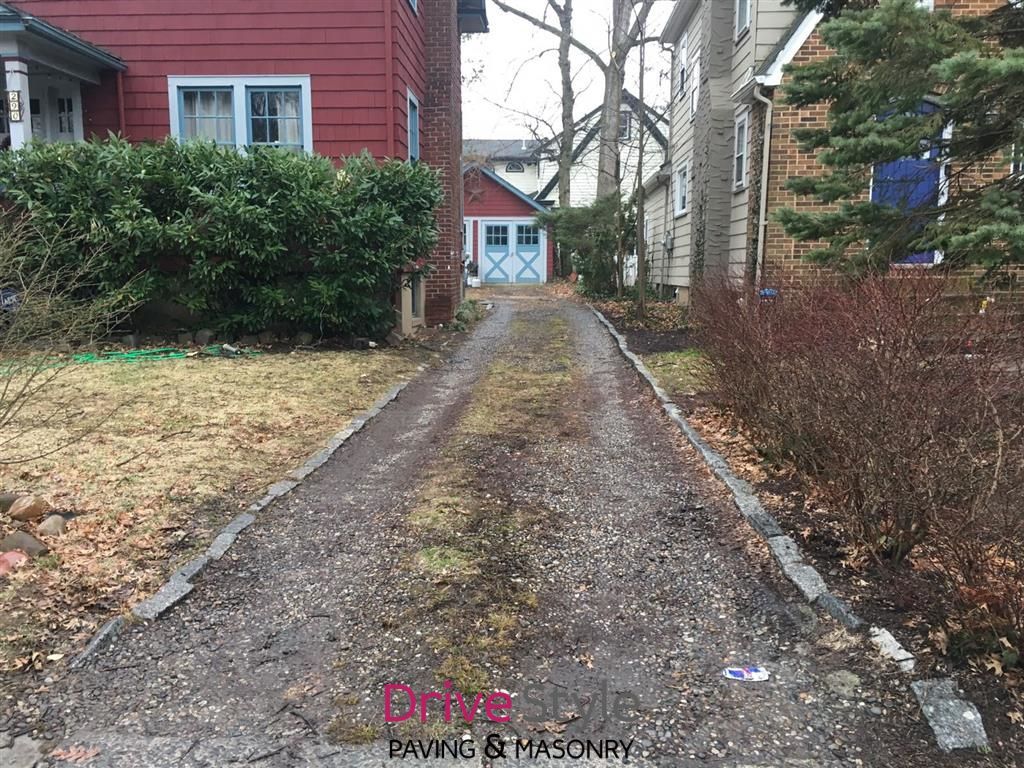 Narrow gravel driveway between red and beige houses, leading to a small garage or shed at the end.