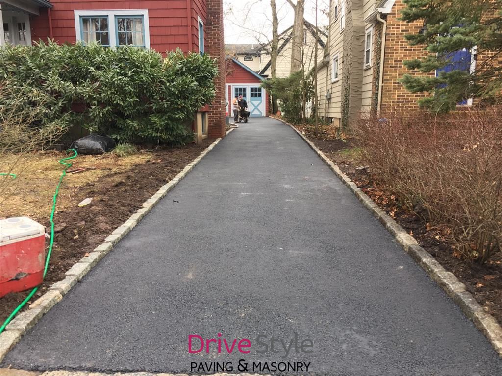 Freshly paved driveway between houses, leading to a backyard gate with a red house on the left and brick wall on the right