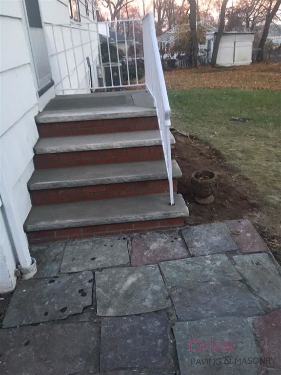 Front steps with white railing leading to a house, beside a stone patio and small grassy yard.