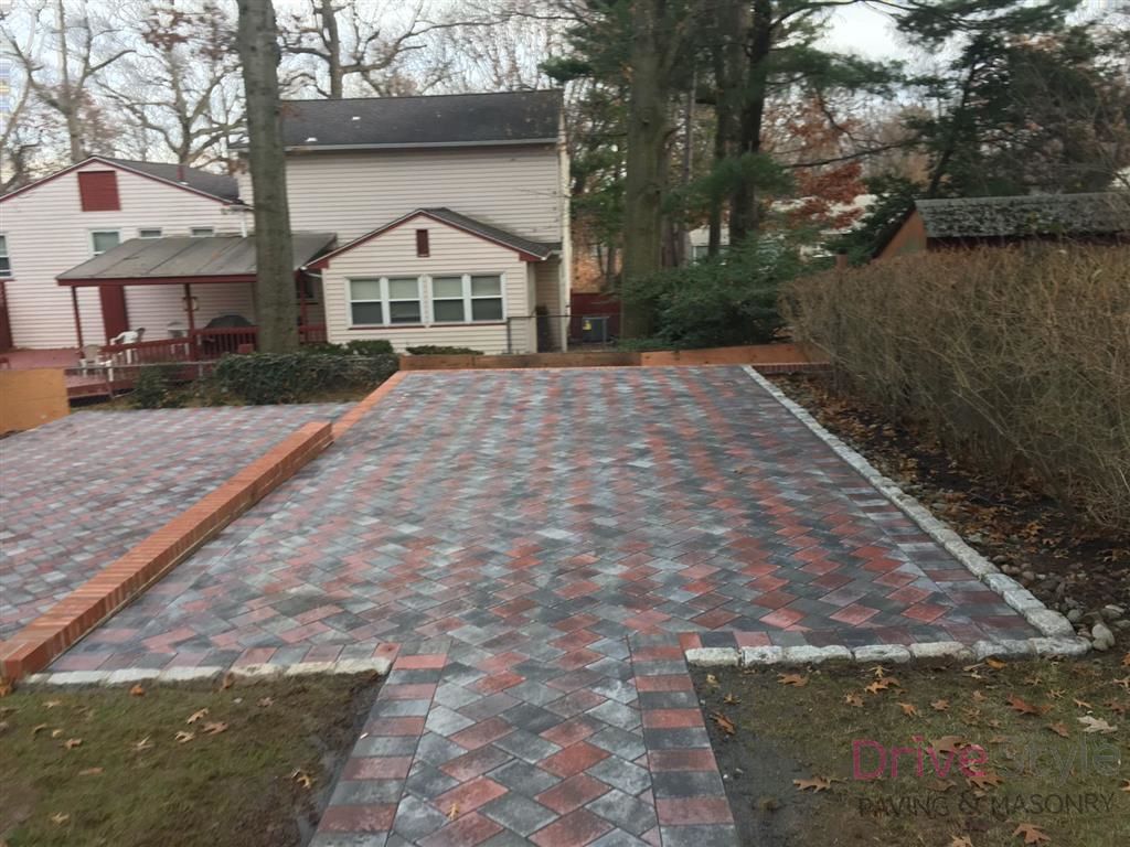 Red and gray brick patio walkway leading to a house in a backyard, bordered by hedges and trees