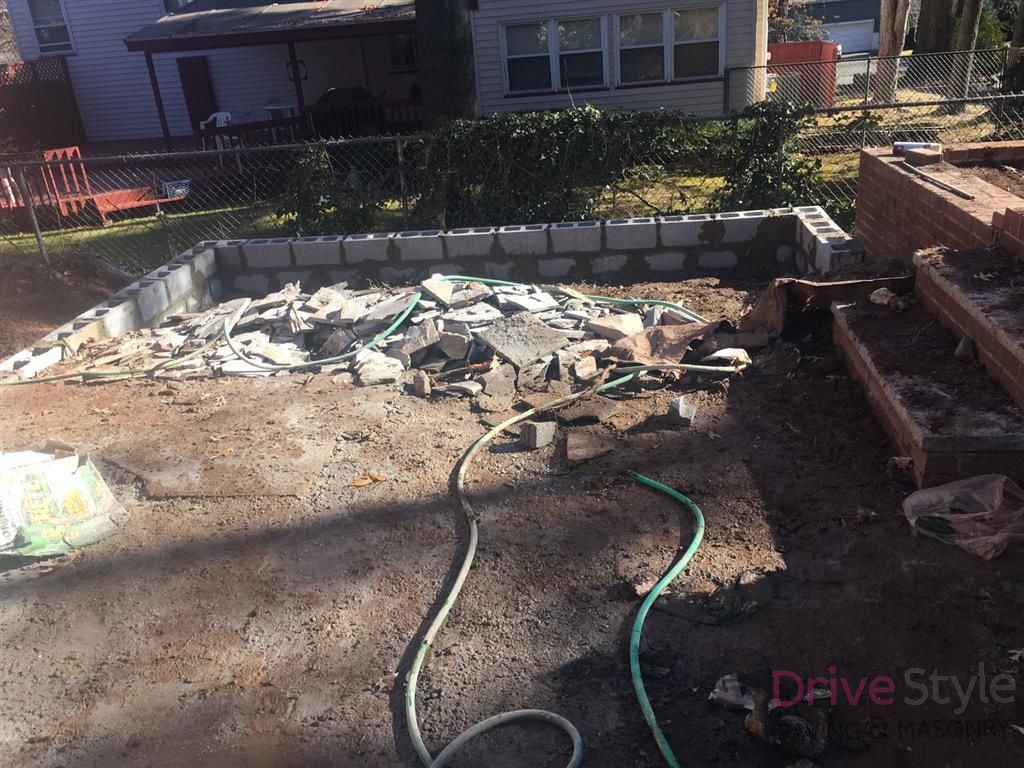 Backyard concrete patio under construction, with rubble, hose, and stacked bricks near a house
