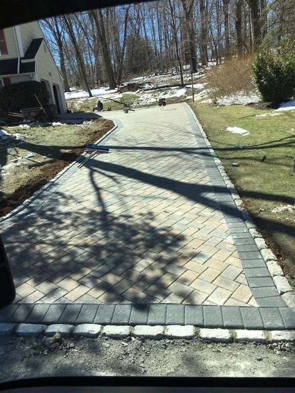 Brick-paver driveway with curved border leading uphill through a wooded suburban area with patches of snow