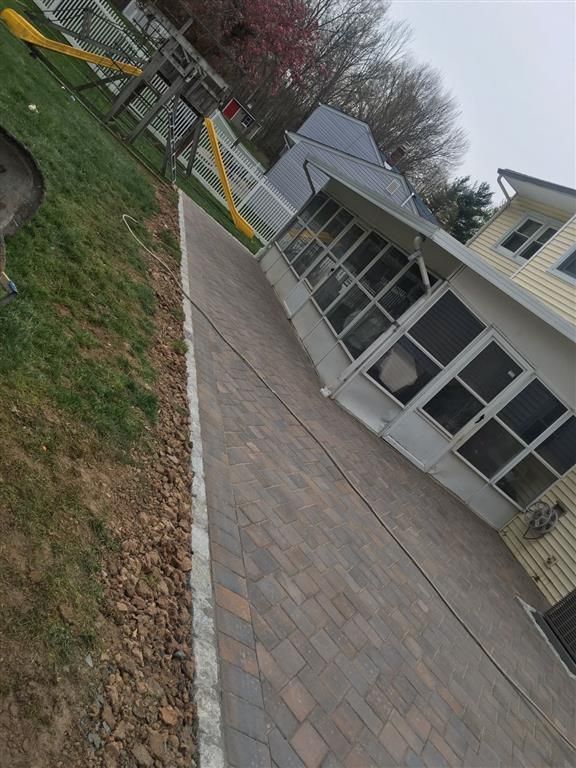Tilted view of a house patio with glass doors, stone pavers, and a grassy yard with trees and a bench.